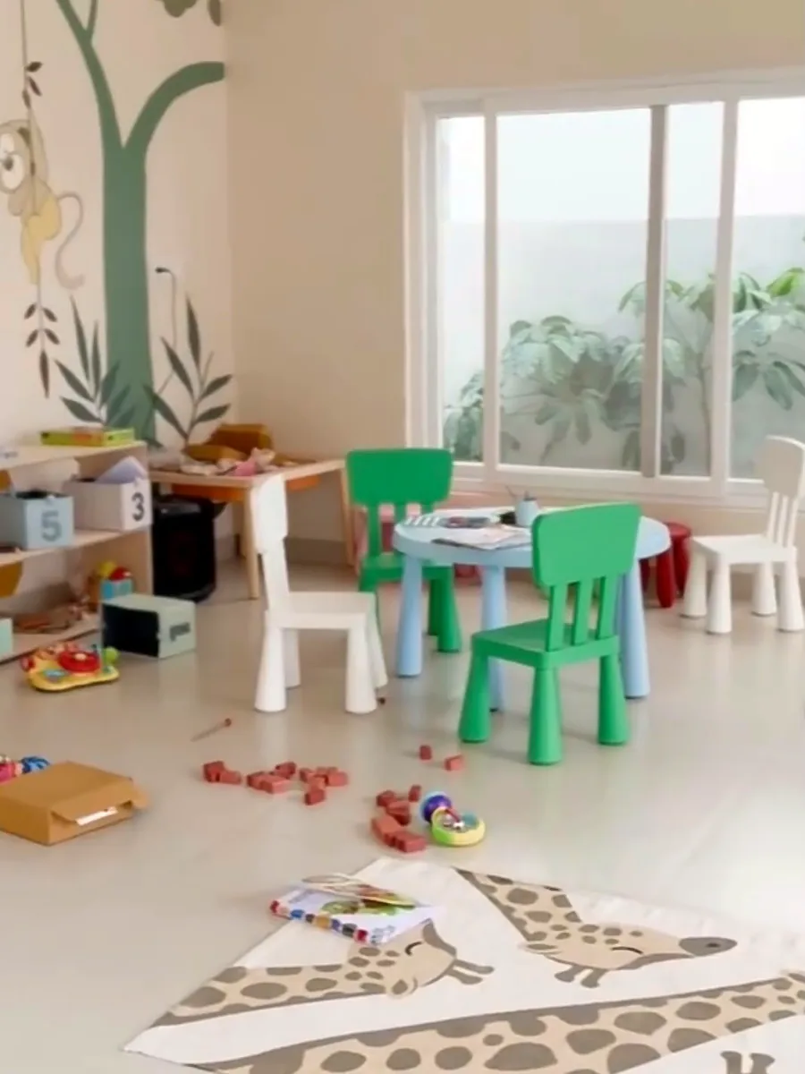 A bright classroom with a tree mural, small green and white chairs, wooden blocks and a giraffe rug